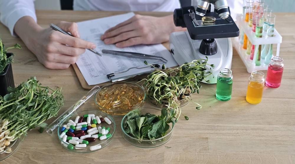 Chercheuse en naturopathie analysant des plantes fraîches, des gélules et des flacons colorés dans un laboratoire avec un microscope.